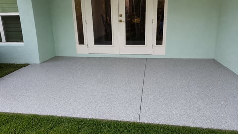 A smooth concrete patio with a center seam, bordered by green grass and a house with white glass doors and light green walls