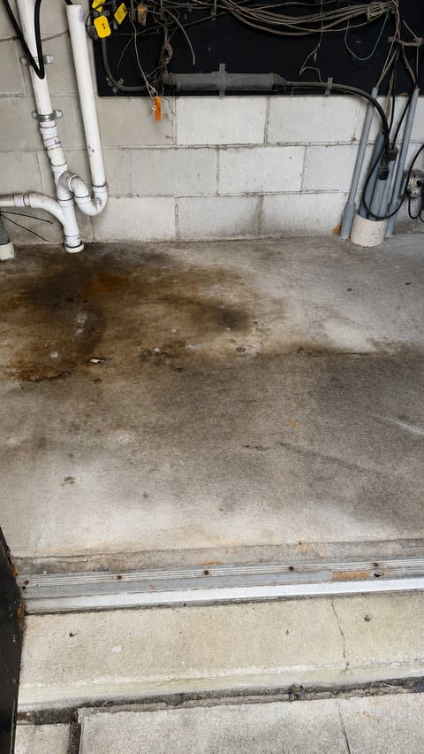 Damp basement floor with wet concrete, PVC pipes on left wall, electrical wiring overhead, and metal beam visible on concrete block wall