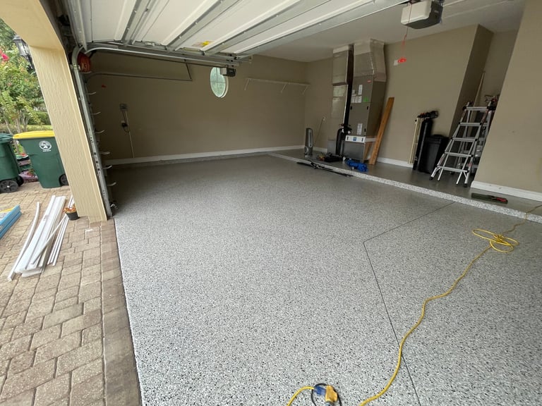 Empty garage interior with gray epoxy floor, white ceiling, storage shelves on right wall, and open garage door showing driveway