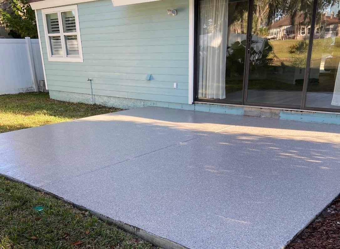 Newly poured concrete patio in front of light blue house with glass sliding doors and shuttered window