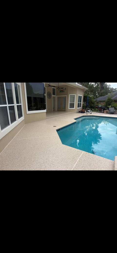 Backyard patio with sparkling blue swimming pool, concrete deck, and beige house with pool area on a sunny day