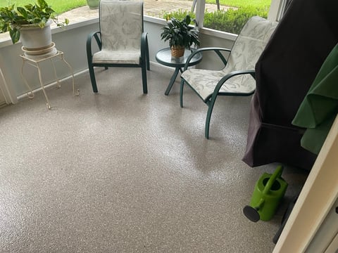 Sunlit sunroom with upholstered chairs, side table with plants, beige carpet flooring, and green watering can in corner.