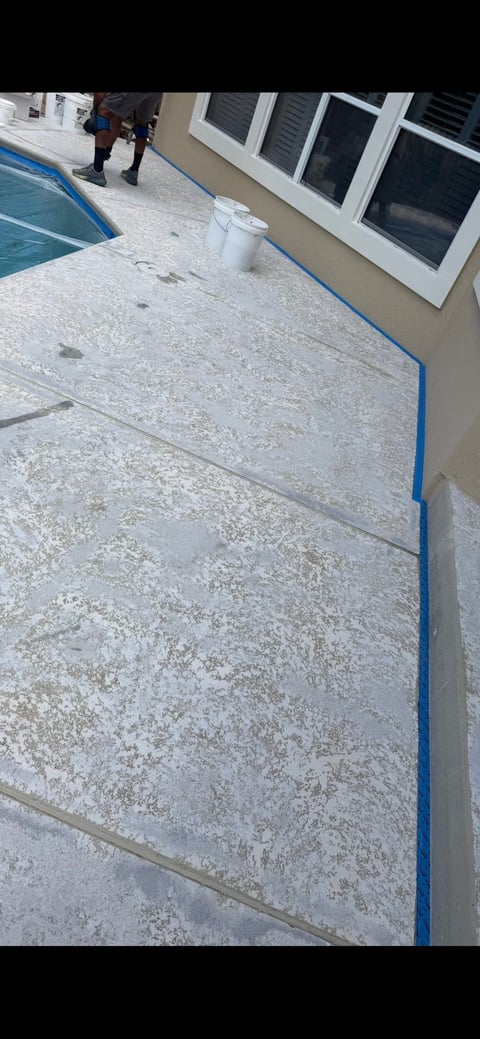Concrete pool deck with blue trim edging, white bucket, and building with shuttered windows in background