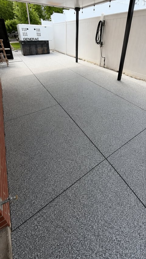 Backyard patio with gray concrete pavers, black pergola, Generac generator, white fence, and coiled electrical cord on wall.