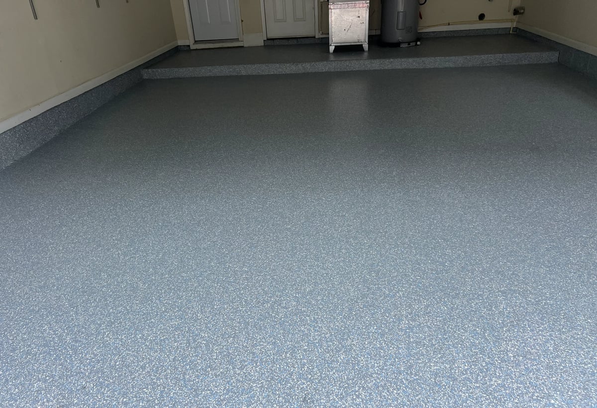 Epoxy-coated garage floor with gray speckled finish and dark border, showing clean empty space with doors and equipment in background